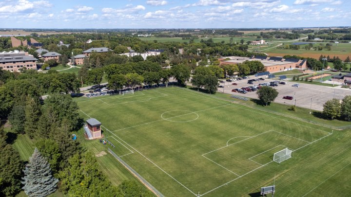 An elevated aerial view of a soccer field surrounded by trees and nearby buildings. The field is neatly lined, with goalposts on both ends and a small building in the corner near a cluster of trees. In the background, several parking lots with parked cars are visible, along with various buildings and open farmland in the distance.